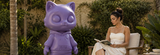 Nina by BANG BANG EG, a purple limited edition art toy sculpture displayed in a luxury garden beside a woman reading, representing contemporary art and collectible design.